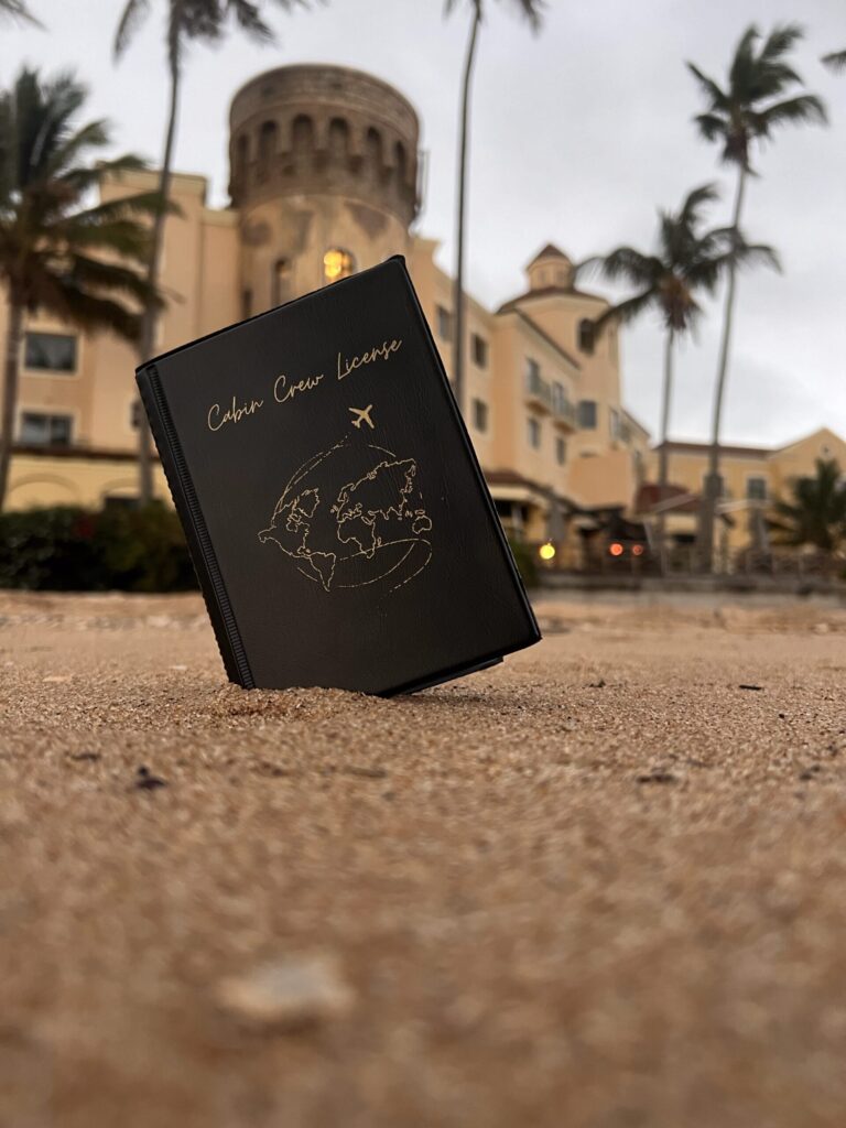 Black Cabin Crew Licence holder with gold foil world map and airplane design photographed on sandy ground with palm trees and a luxury resort in the backgorund