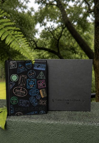 Cabin Crew License Book with colorful travel stamp design displayed beside a Cabin Crew Logbook outdoors in a lush green nature setting.